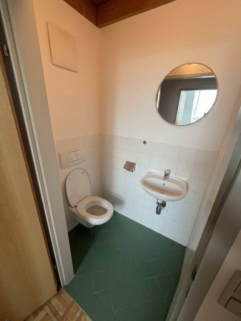 Kleines Gäste-WC mit Toilette, Handwaschbecken, runden Spiegel und grünen Bodenfliesen.