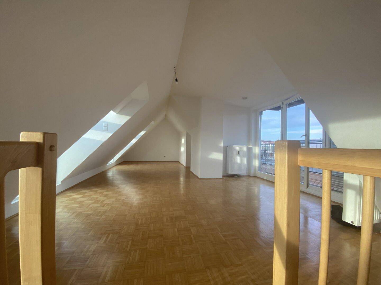 Helles Dachgeschoss-Wohnzimmer mit Parkettboden, schrägen Wänden und Zugang zu einem Balkon.