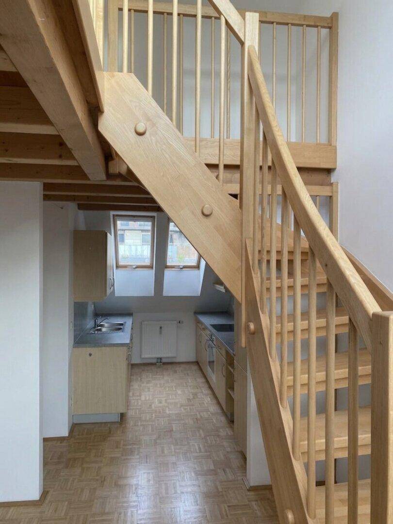 Holztreppe führt zu einem oberen Bereich mit Blick auf die Küche und Holzbalken.