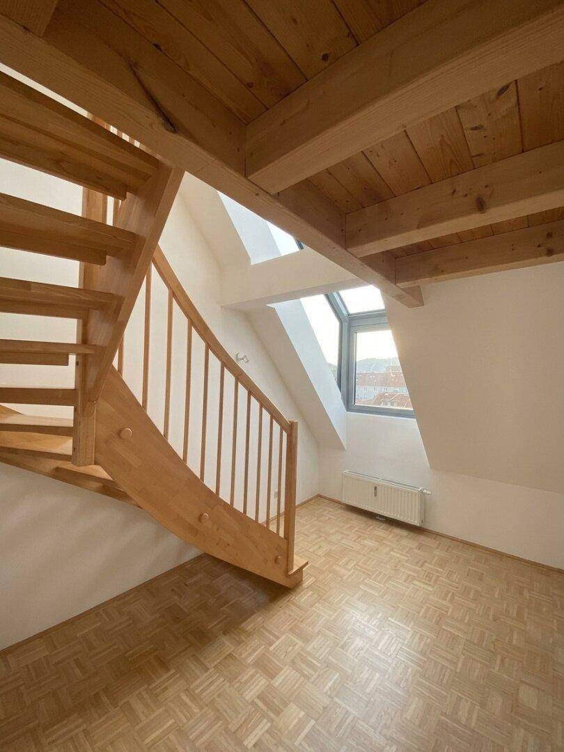 Holztreppe in einem hellen Bereich mit sichtbaren Holzbalken und einem Dachfenster.