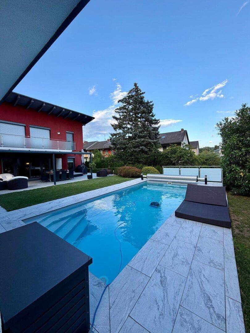Blauer Swimmingpool im gepflegten Gartenbereich mit Liegefläche und rotem Haus im Hintergrund.