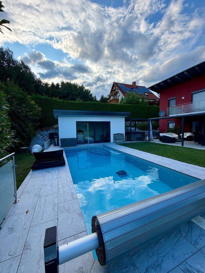 Großer Swimmingpool im Garten mit gefliestem Bereich und Blick auf das moderne Poolhaus.