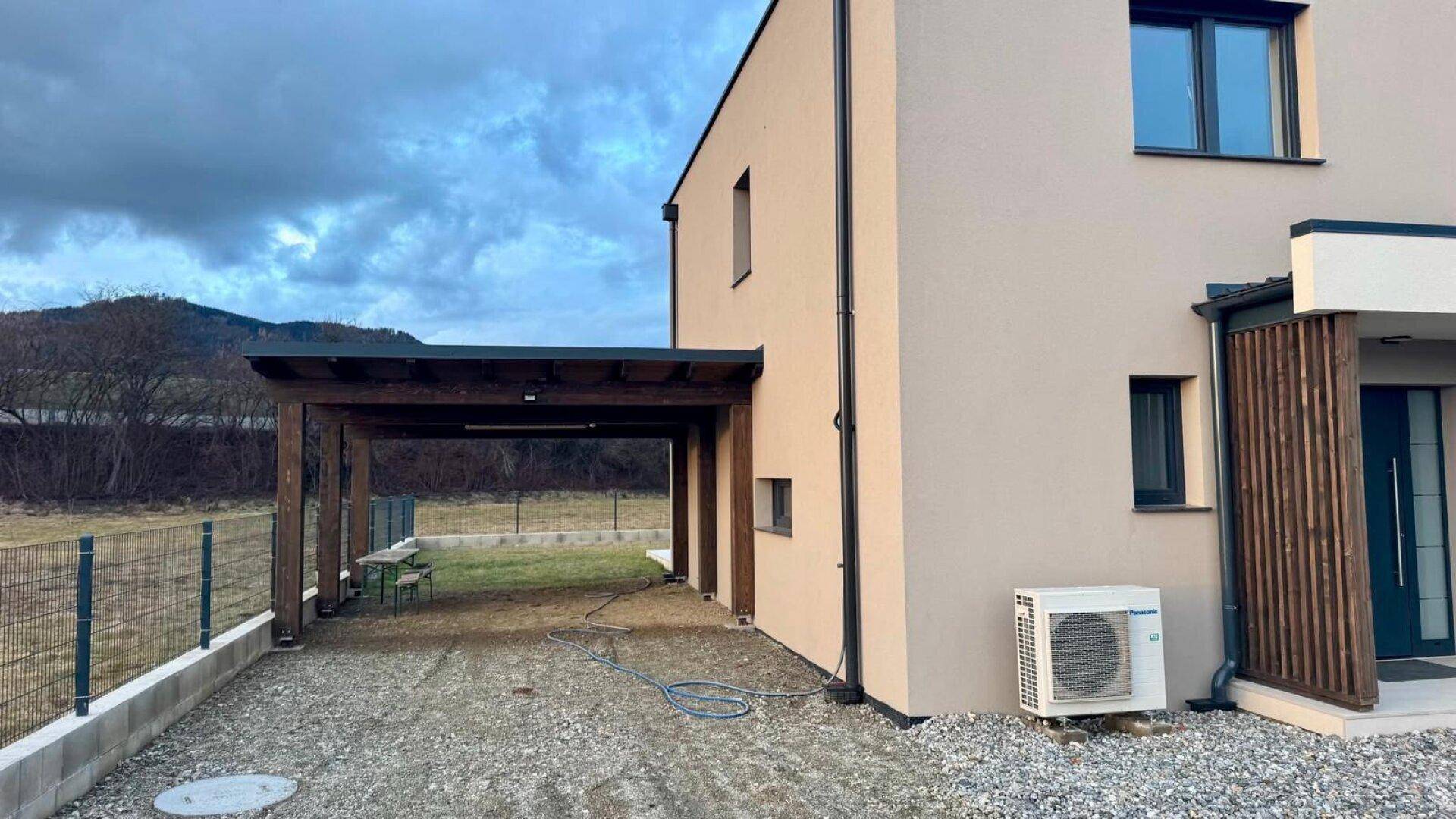 Seitenansicht des Hauses mit Holz-Carport und Kiesauffahrt, daneben eine Klimaanlage.