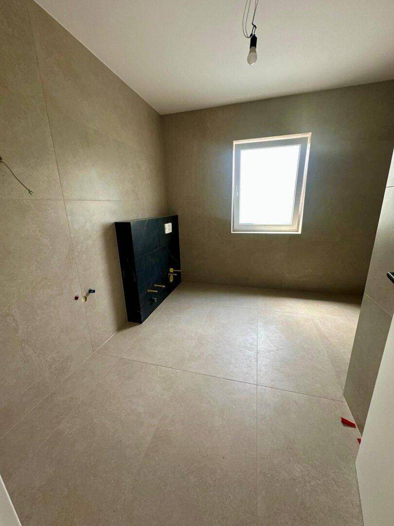 Geräumiges Badezimmer mit beigefarbenen Großformatfliesen, Fenster und vorbereiteter Toiletteninstallation.