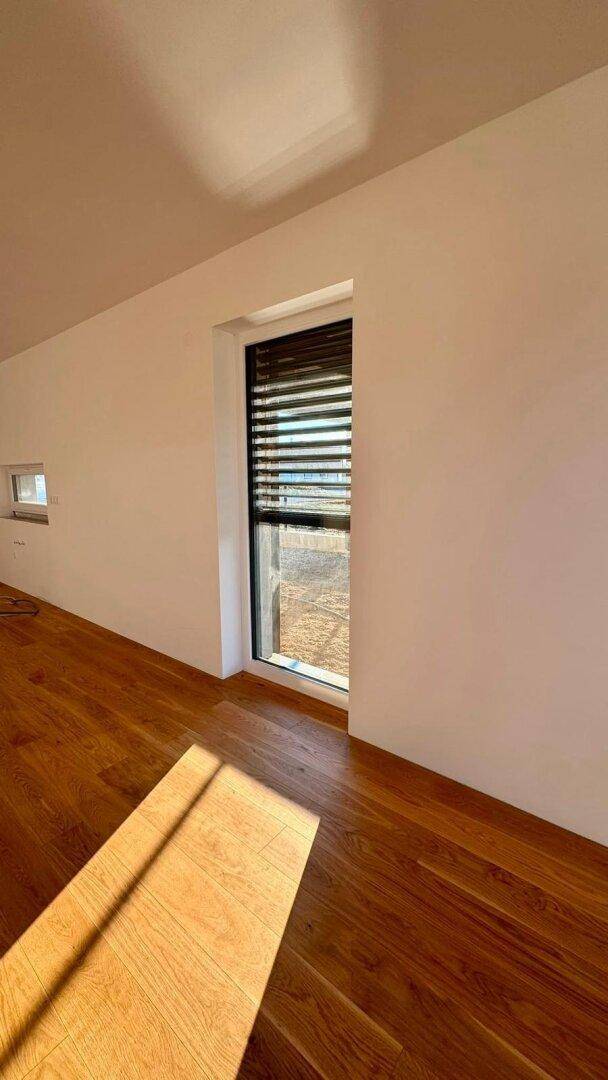 Heller Raum mit Holzboden und hohem Fenster mit Jalousien, das viel Tageslicht hereinlässt.