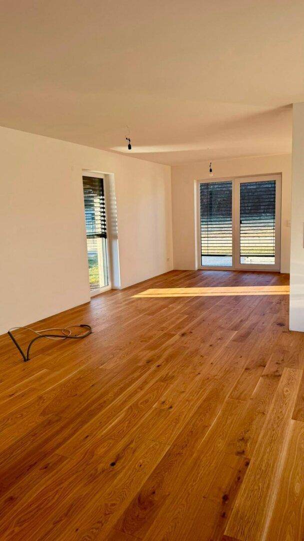 Geräumiger, heller Raum mit Holzboden und zwei großen Fenstern mit Jalousien.