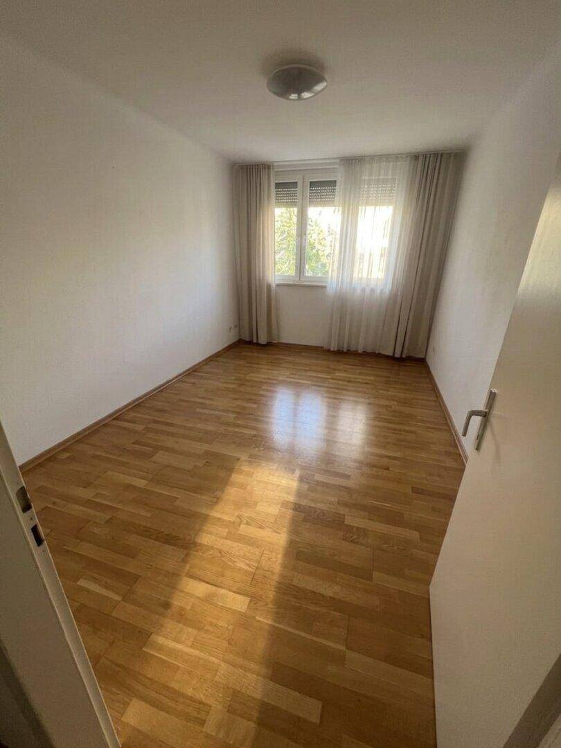 Leeres Zimmer mit hellem Parkettboden und großem Fenster, ideal als Schlaf- oder Arbeitszimmer.