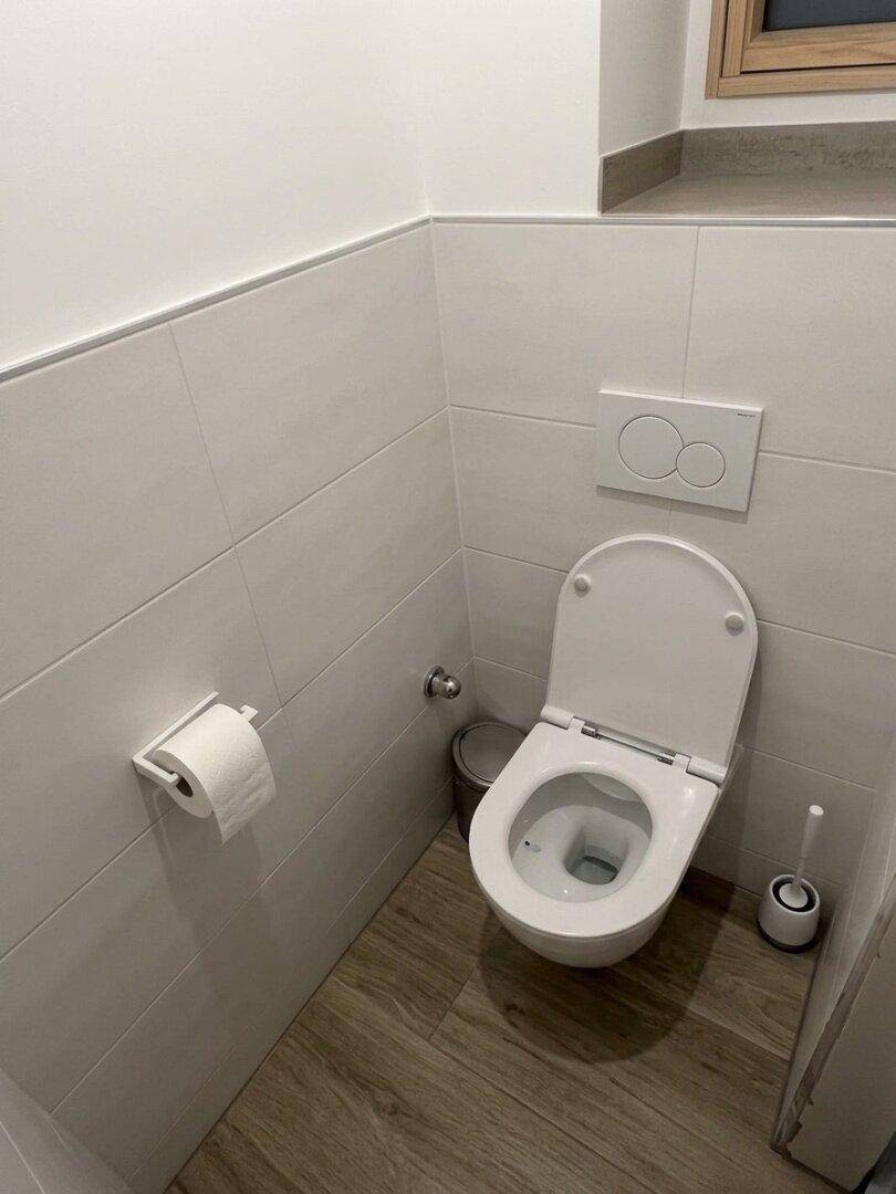 Separates WC mit wandhängender Toilette, kleinem Fenster und hellen Fliesen.