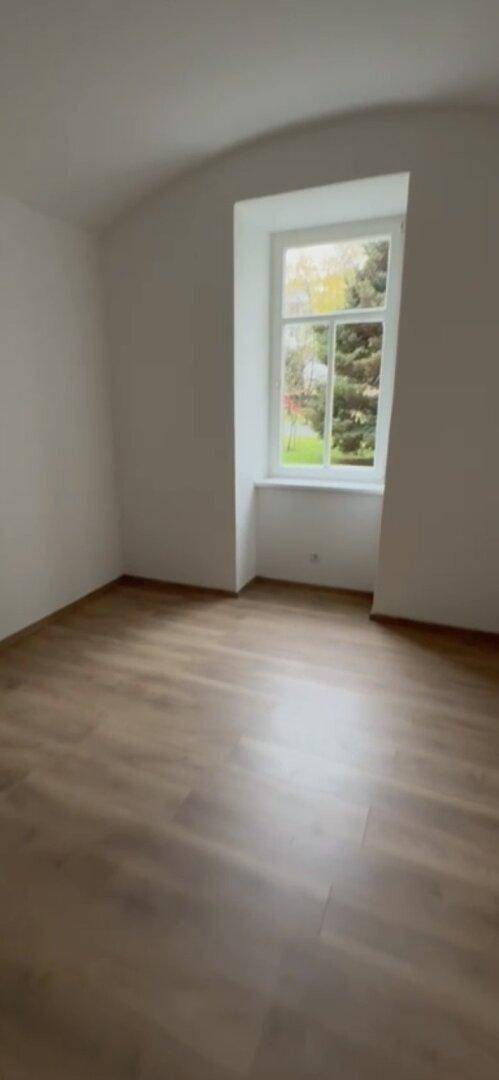 Heller, leerer Raum mit Laminatboden und einem Fenster mit Blick ins Grüne.