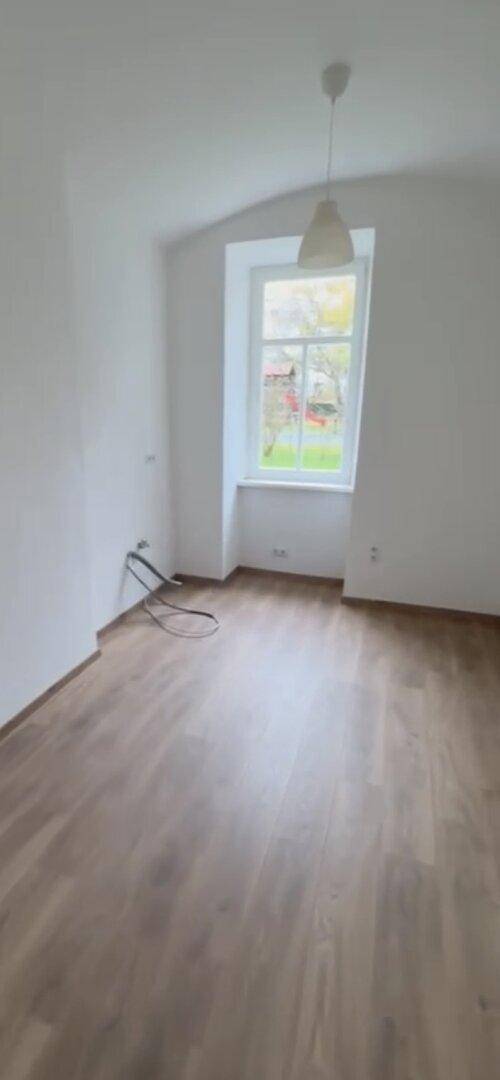 Leerer Raum mit Laminatboden, einem Fenster und einer einfachen Deckenleuchte.