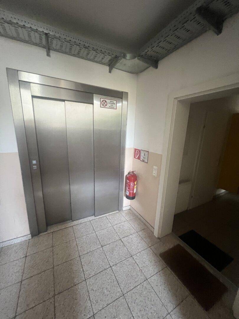 Moderner Aufzug in einem Gemeinschaftsbereich mit Fliesenboden und Brandschutzvorrichtung.
