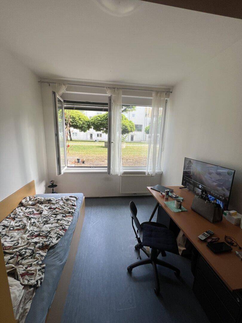 Helles Schlafzimmer mit Einzelbett, Schreibtisch und Blick ins Grüne durch das Fenster.