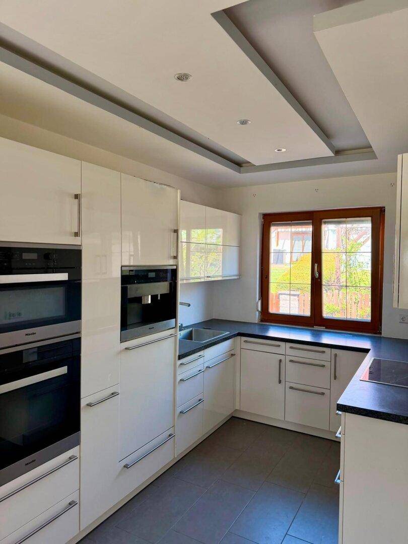 Moderne Küche mit weißen Hochglanzfronten, Einbaugeräten und Fenster zum Gartenblick.