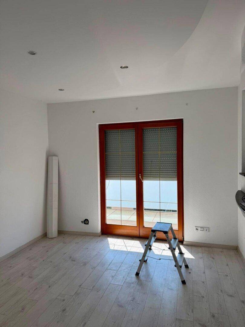 Heller, leerer Raum mit hellem Laminatboden und großen Fenstertüren zum Außenbereich.