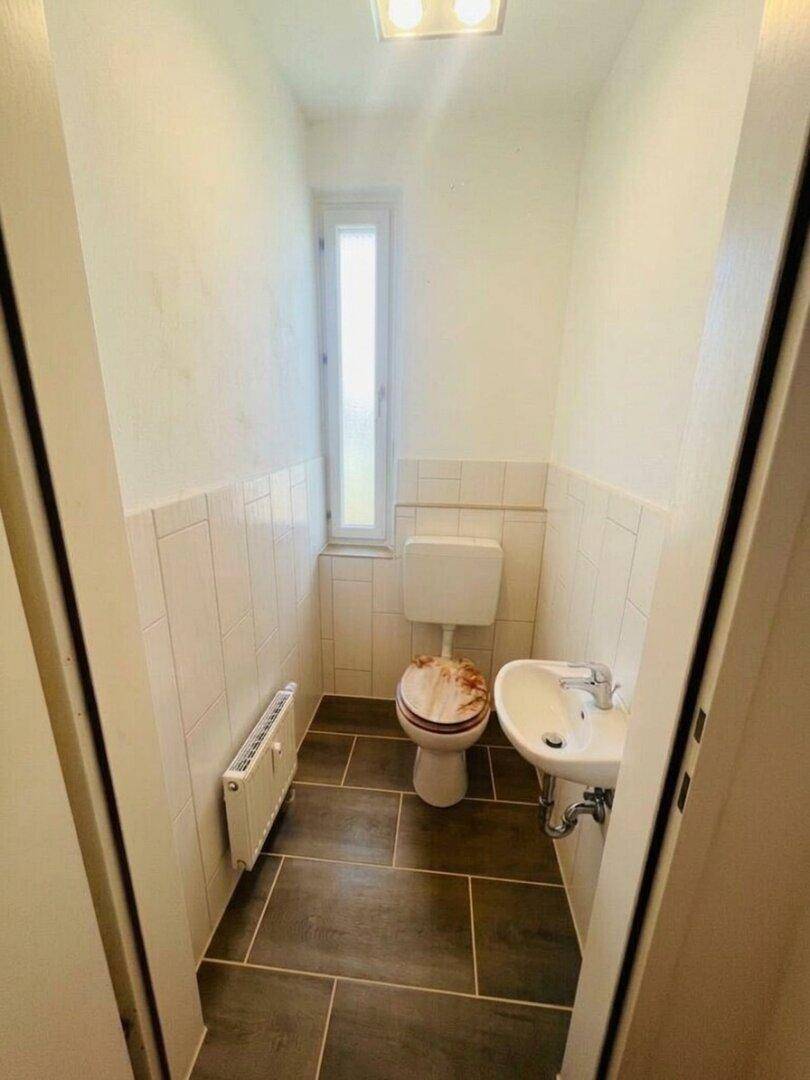 Kleines Gäste-WC mit Fenster, Wand-WC und Einzelwaschbecken, gefliest mit dunklen Bodenfliesen.