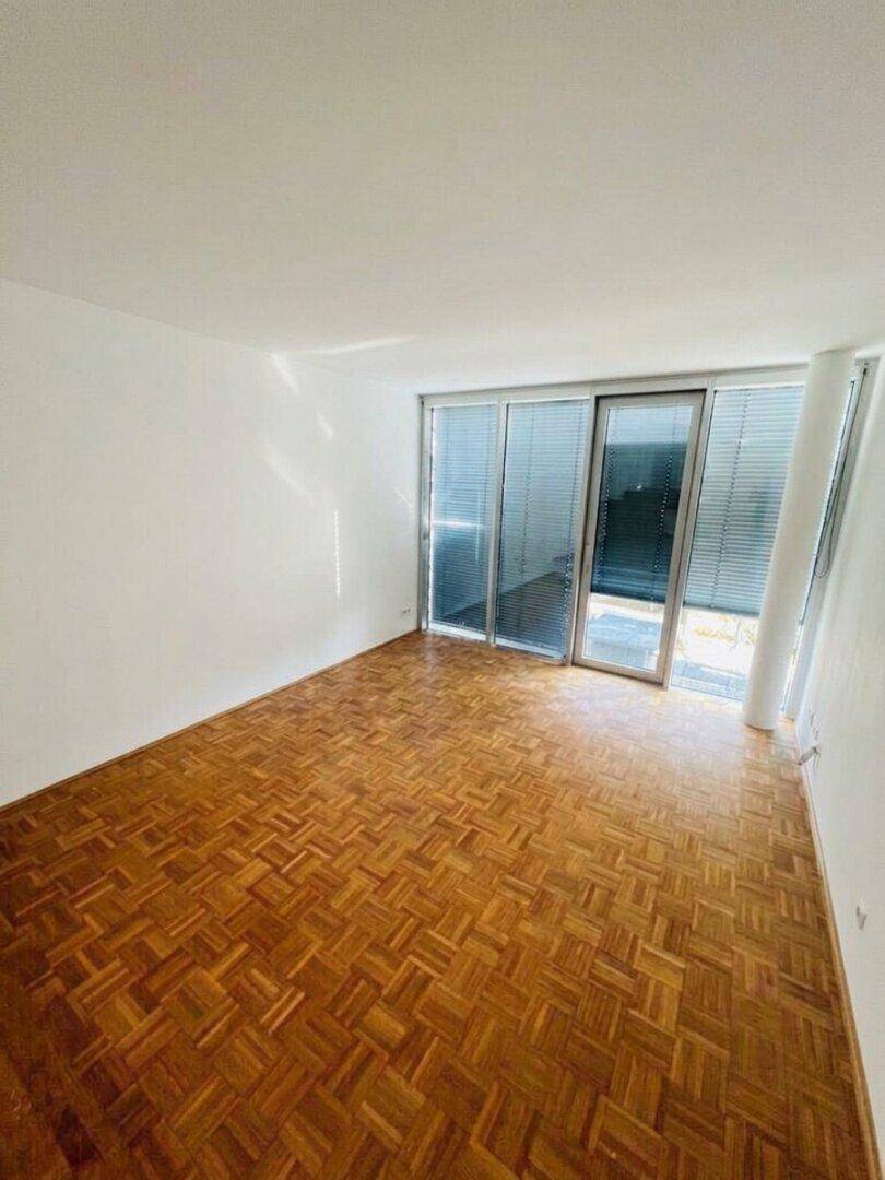 Geräumiges, leeres Zimmer mit Parkettboden und großen Fenstern, die viel Tageslicht hereinlassen.