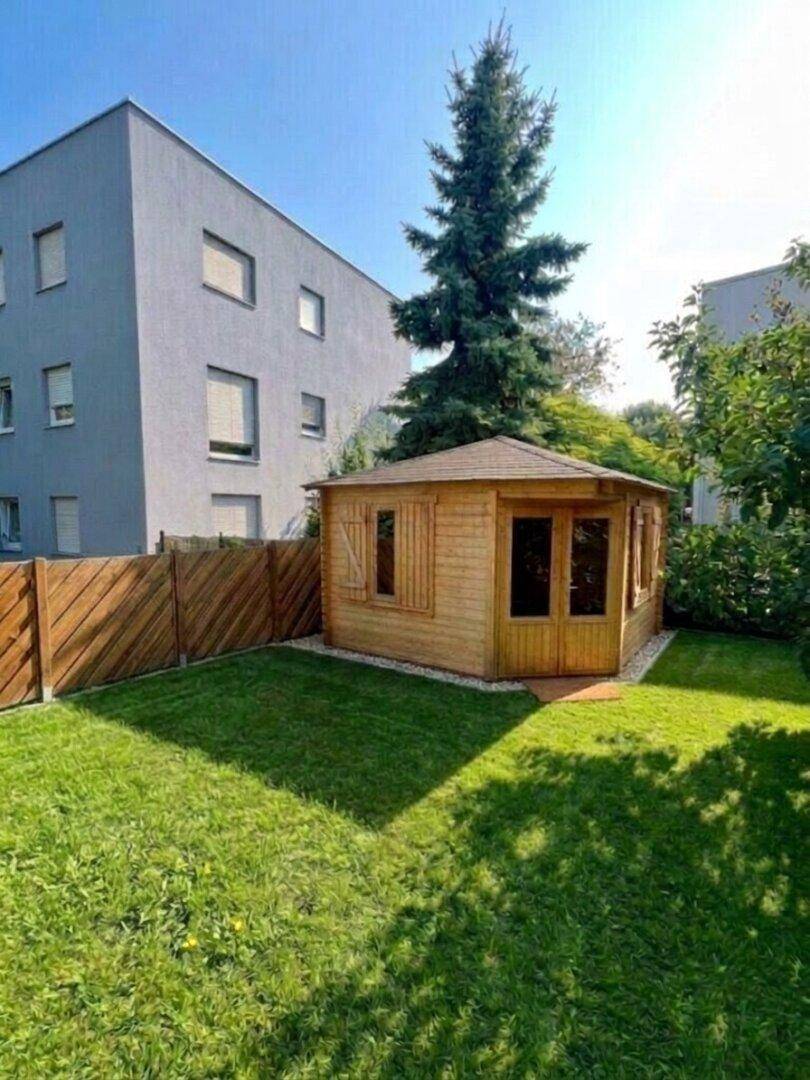 Gepflegter Garten mit Rasenfläche und einem charmanten Holzgartenhaus unter einem großen Baum.