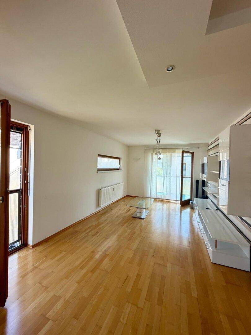 Helles, leeres Wohnzimmer mit Parkettboden, weißen Wänden und Zugang zum Balkon.