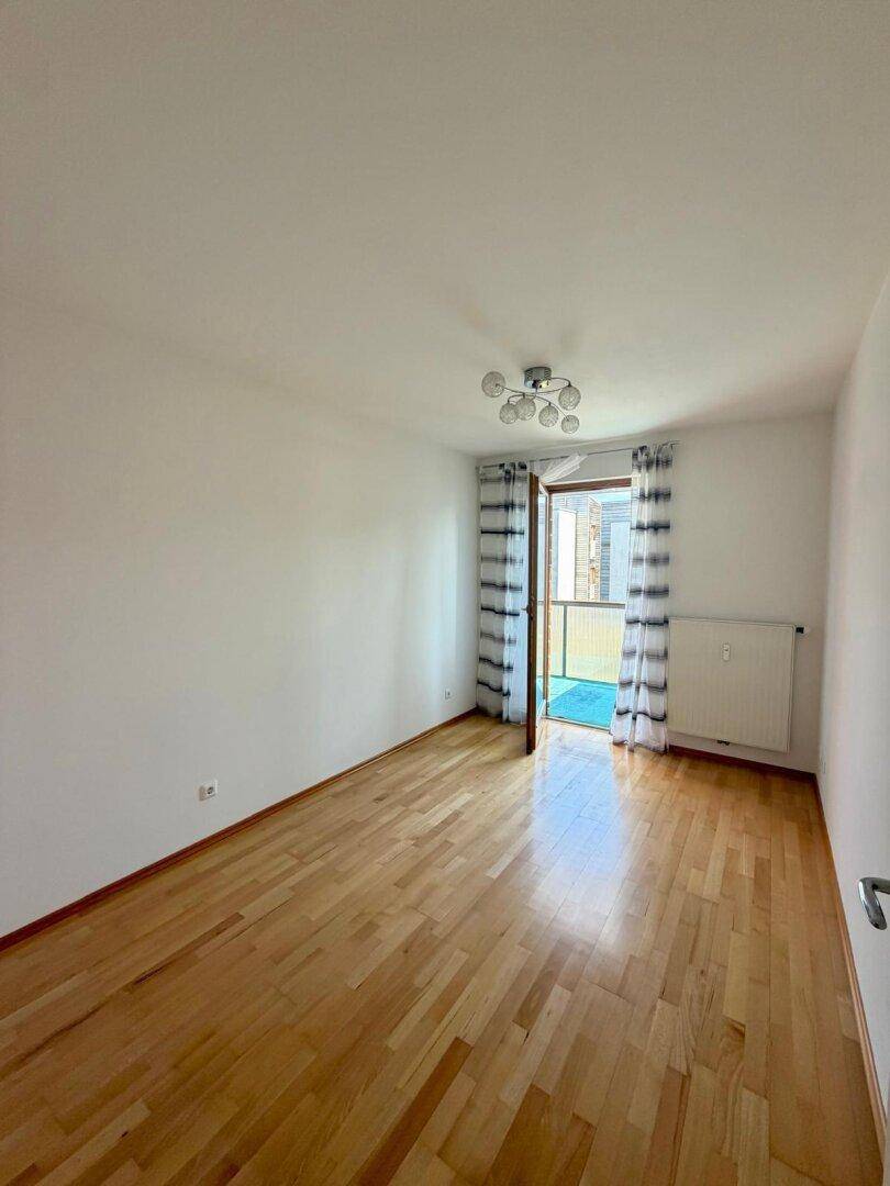 Leeres Schlafzimmer mit Parkettboden, weißen Wänden und Zugang zum Balkon.