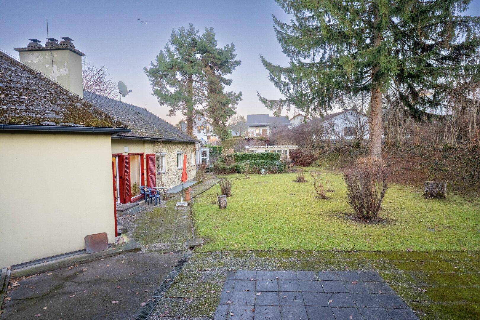 Weitläufiger Garten mit Rasenfläche und gepflastertem Bereich, umgeben von Bäumen.