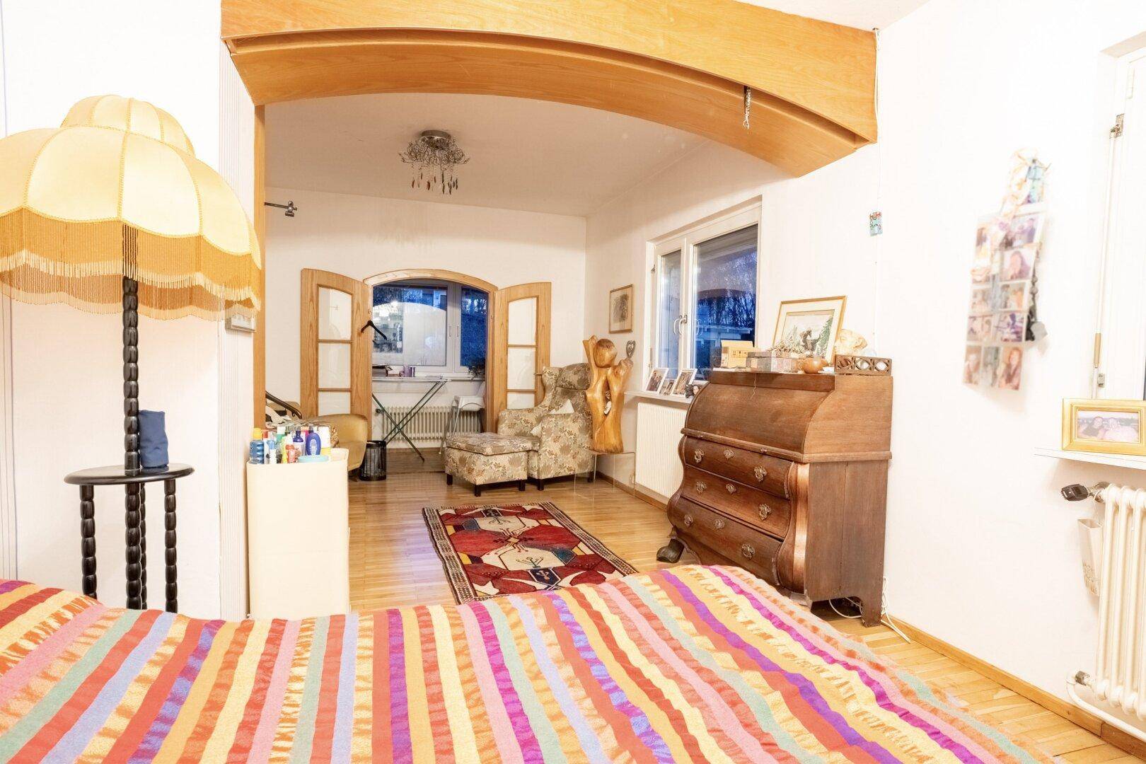 Helles Schlafzimmer mit Holzboden und Blick in den angrenzenden Wohnbereich mit Fenstern.