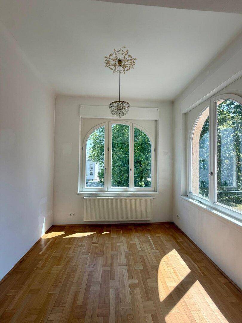 Heller Raum mit Fischgrätparkett und großen, bogenförmigen Fenstern mit Blick ins Grüne.