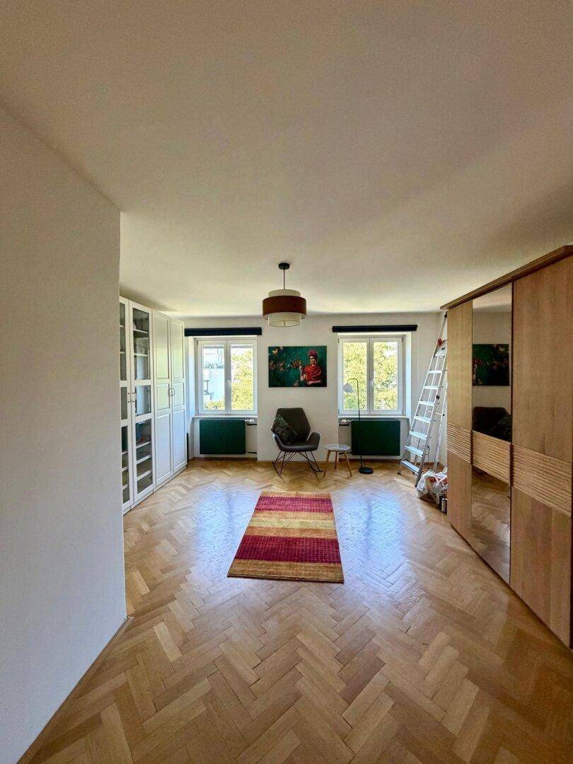 Schlafzimmer mit Fischgrätparkett, Einbauschränken und zwei Fenstern mit Blick ins Grüne.