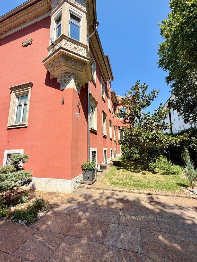 Eckansicht der Villa mit gepflastertem Bereich und Bäumen, die Schatten spenden.