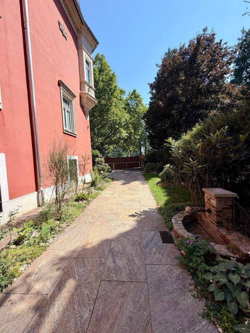 Gepflasterter Weg entlang der Villa mit Bepflanzung und einem kleinen dekorativen Brunnen.