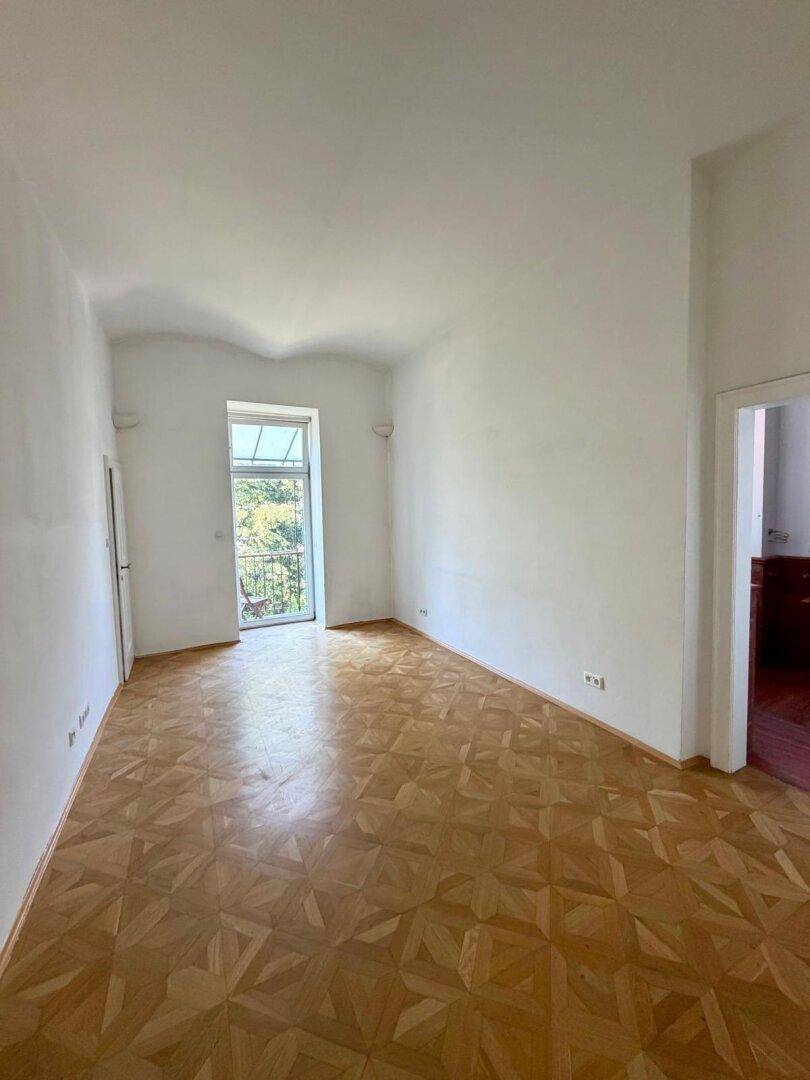 Helles Zimmer mit Fischgrätparkett und Zugang zu einem kleinen Balkon.