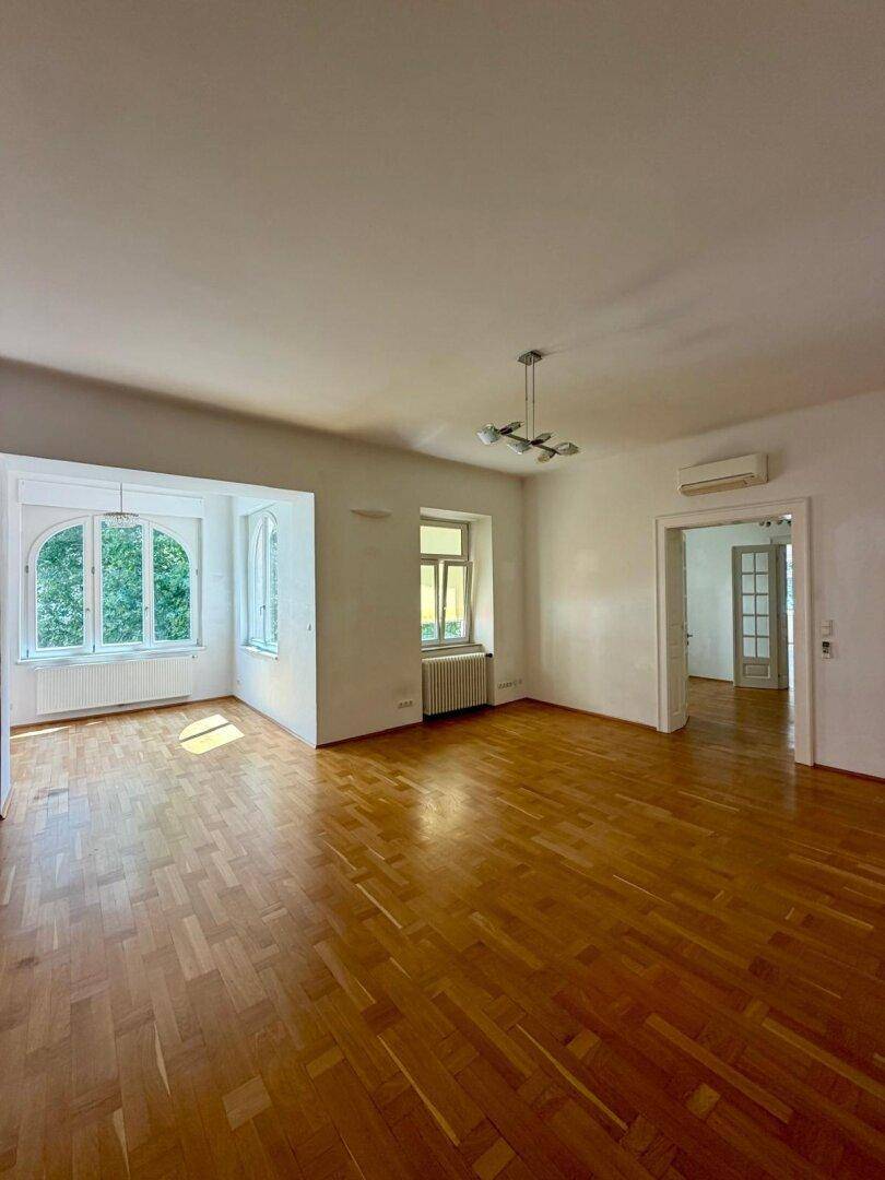 Helles Wohnzimmer mit Parkettboden, hohen Decken und großen Fenstern.