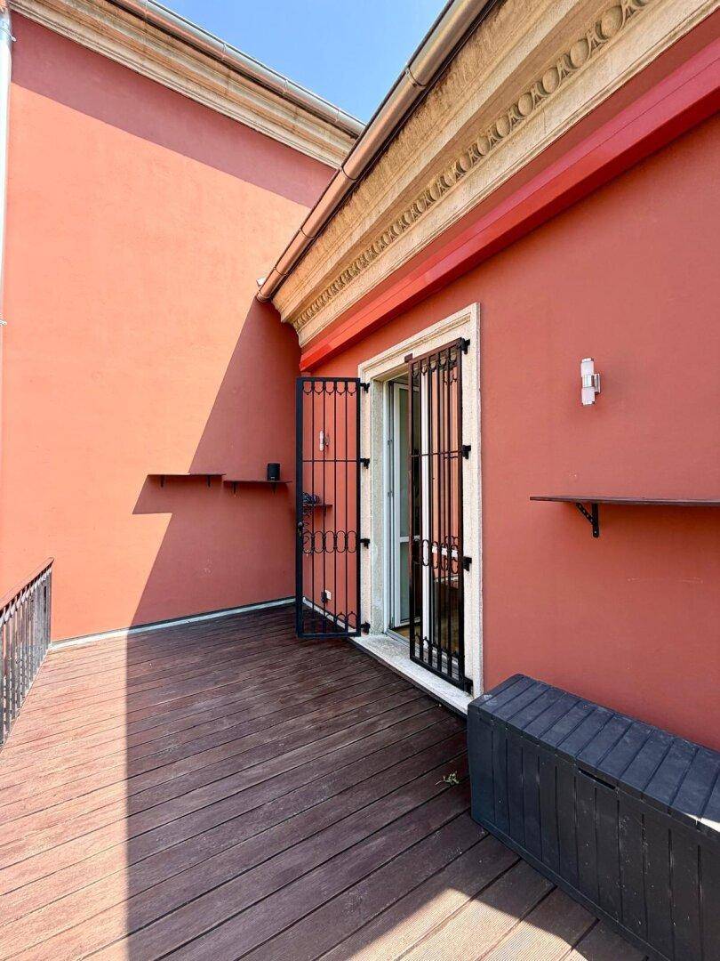 Sonniger Balkon mit Holzdeck, roter Fassade und schmiedeeisernem Geländer.