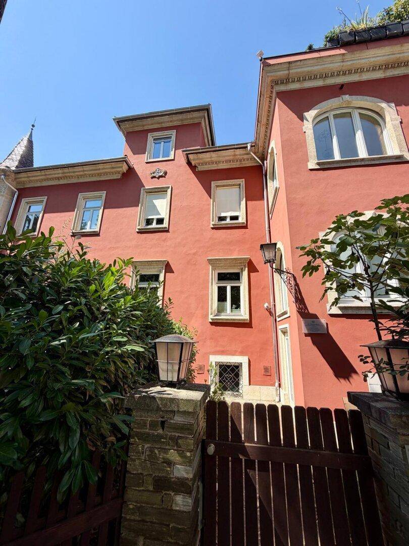 Rote Gebäudefassade mit traditionellen Fenstern, Zaun und üppiger Bepflanzung.