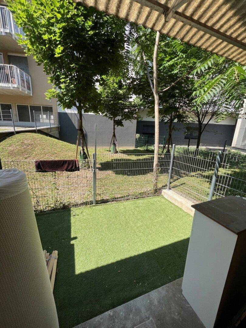 Kleiner eingezäunter Gartenbereich mit Kunstrasen und Bäumen im Hintergrund.
