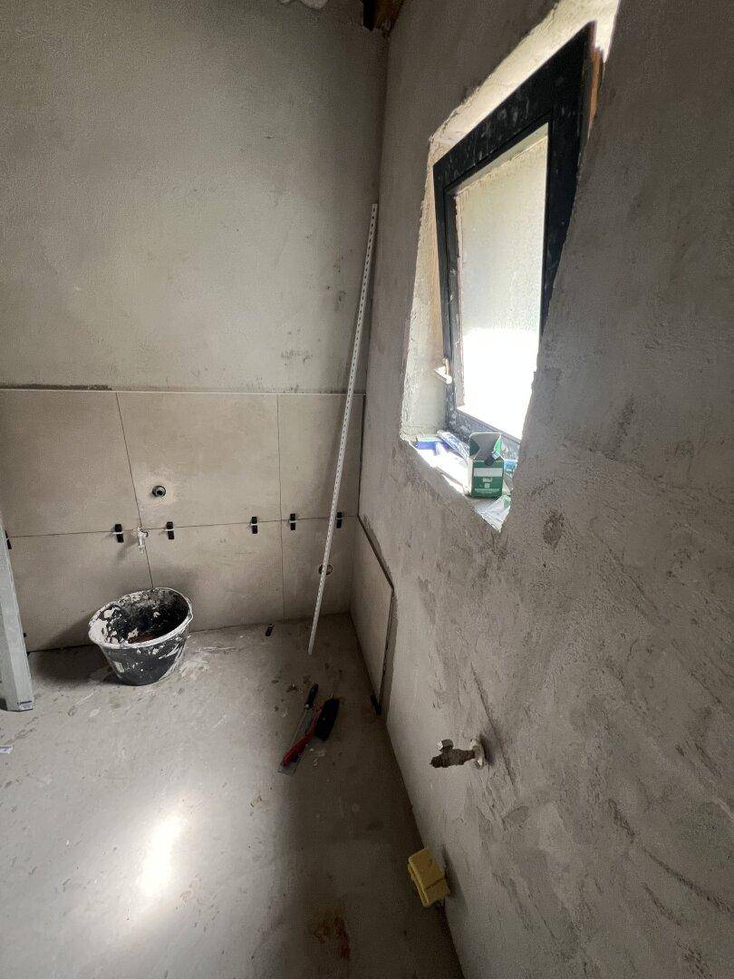 Badezimmer im Rohbau mit Fenster und teilweise gefliesten Wänden, viel natürliches Licht.