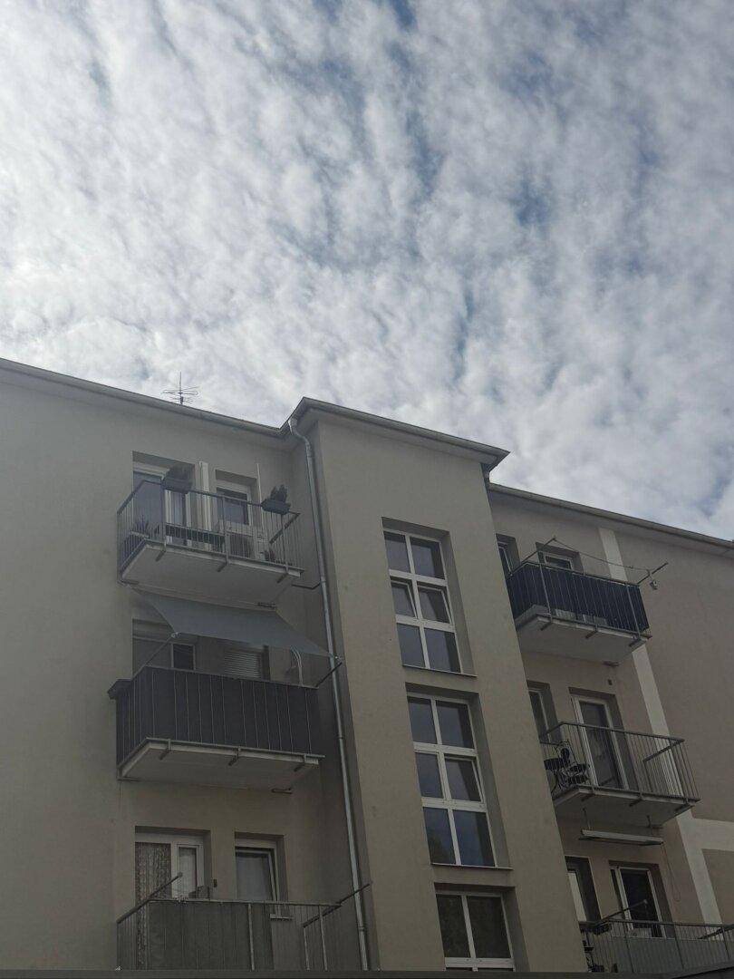 Fassade eines modernen Mehrfamilienhauses mit mehreren Balkonen und großen Fenstern unter bewölktem Himmel.