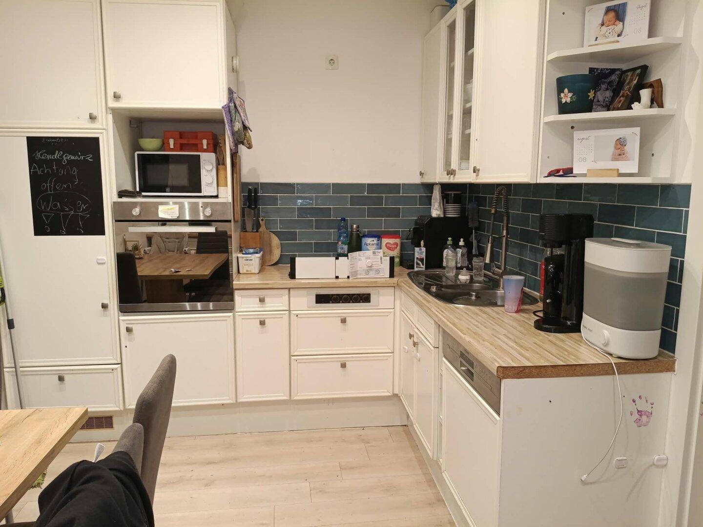 Funktionale Küche mit weißen Schränken, Holztheke, blauen Fliesen und integrierten Geräten.
