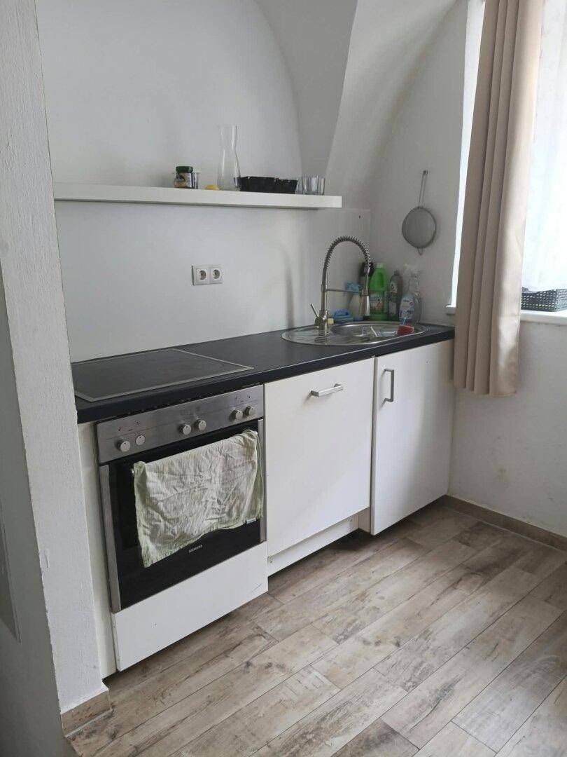 Kleine Küchenzeile mit Kochfeld, Backofen und Spüle unter einem Dachschrägenfenster.