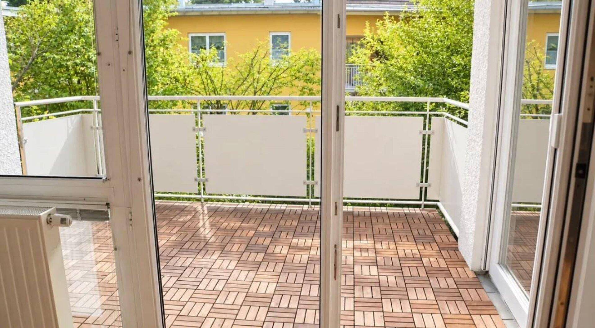 Blick von einem Innenraum auf einen Balkon mit Holzdielen und grüner Umgebung.