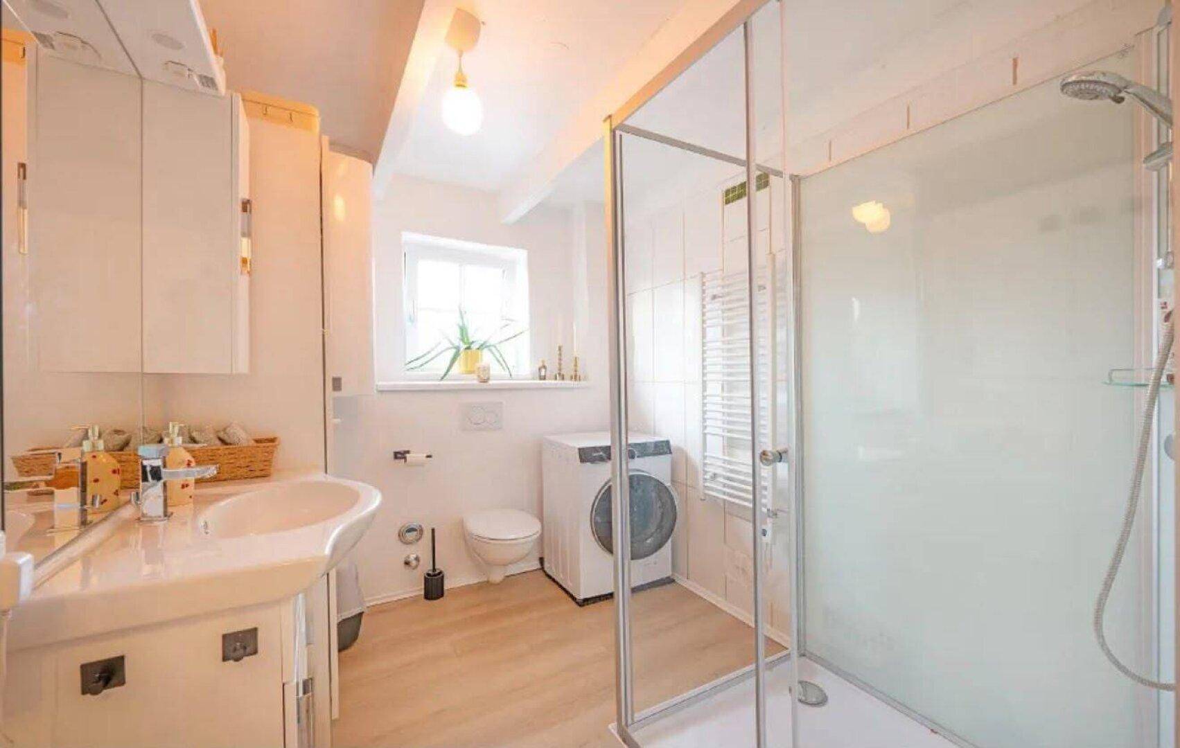 Helles Badezimmer mit Duschkabine, Waschbecken, WC und integrierter Waschmaschine.