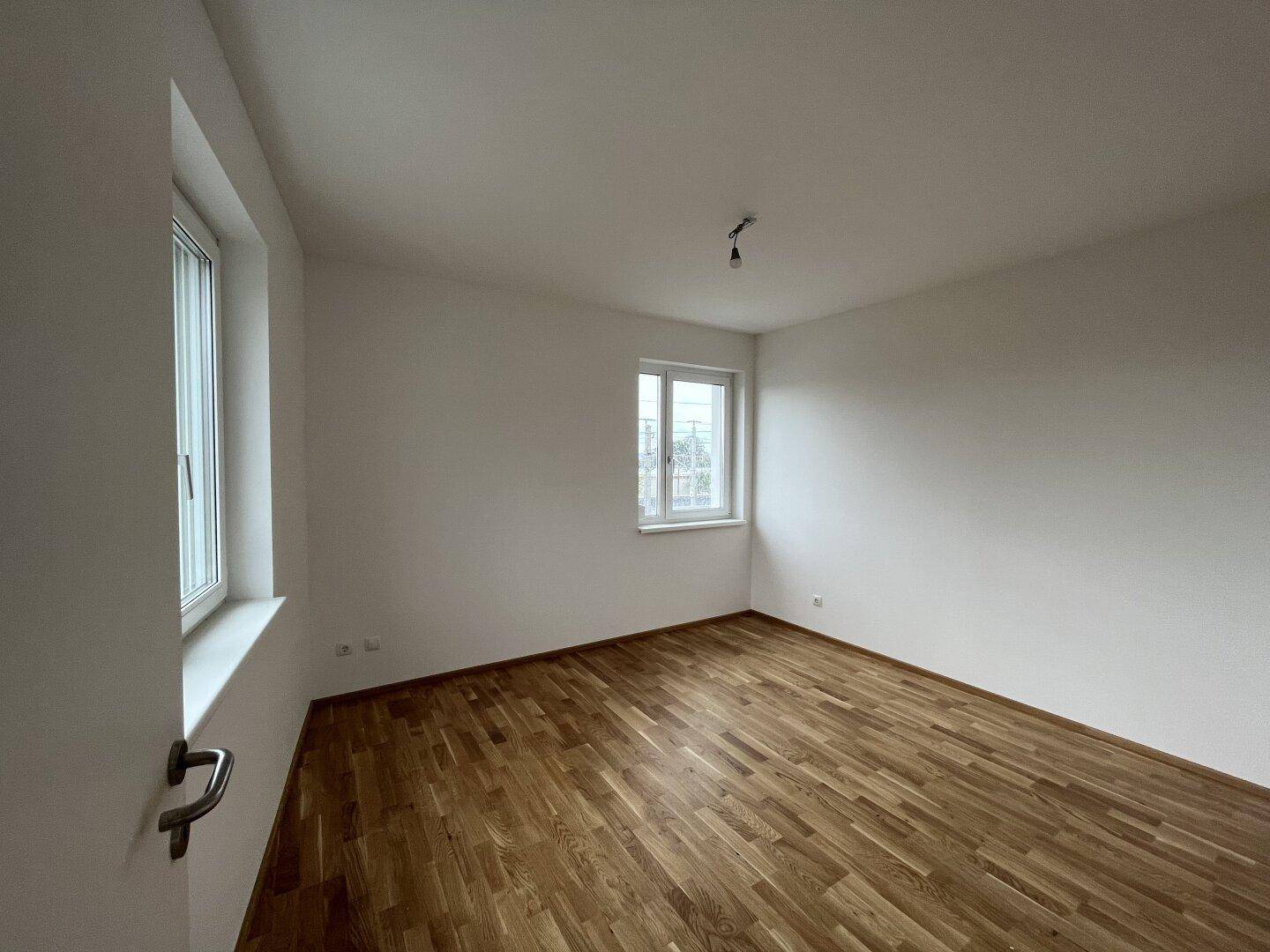 Leeres Schlafzimmer mit hellem Parkettboden und zwei Fenstern für viel Tageslicht.
