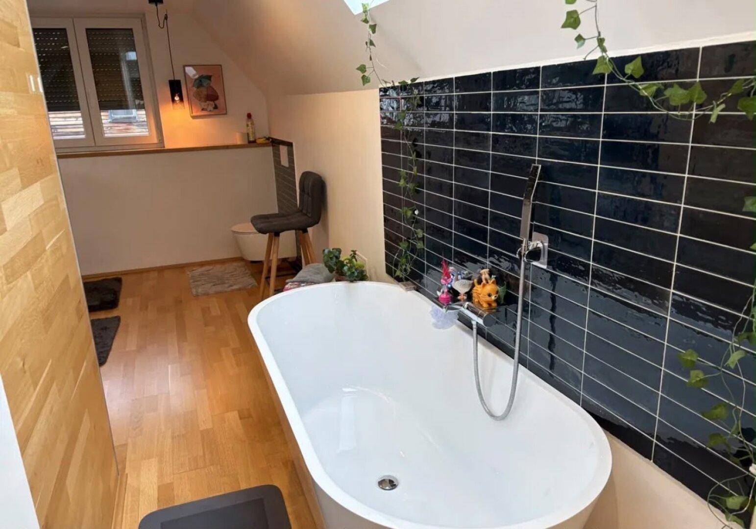 Modernes Badezimmer im Dachgeschoss mit freistehender Badewanne und dunklen Wandfliesen.