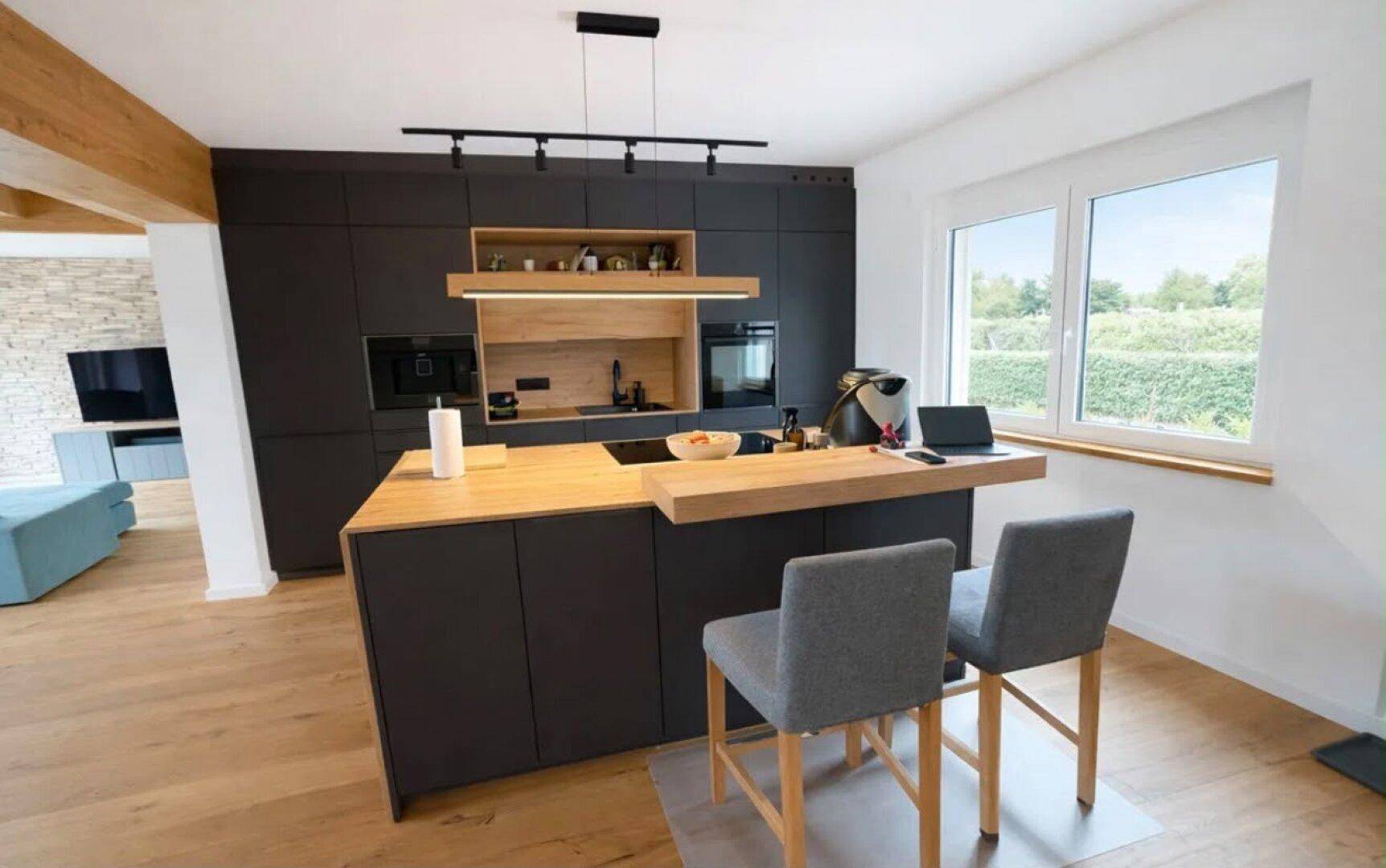 Moderne offene Küche mit dunklen Schränken, Holztheke und Blick ins Grüne durch große Fenster.
