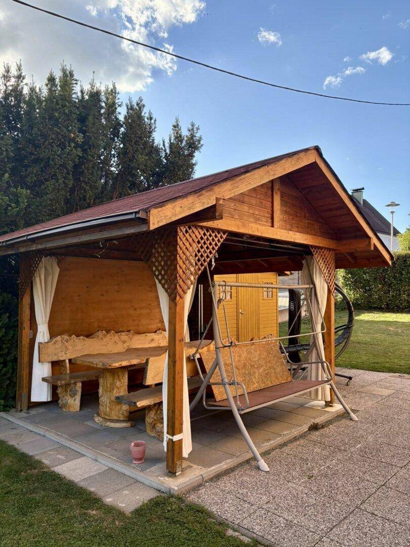 Rustikaler Holzpavillon im Garten mit Schaukel und massiver Holzsitzgruppe für gemütliche Stunden.