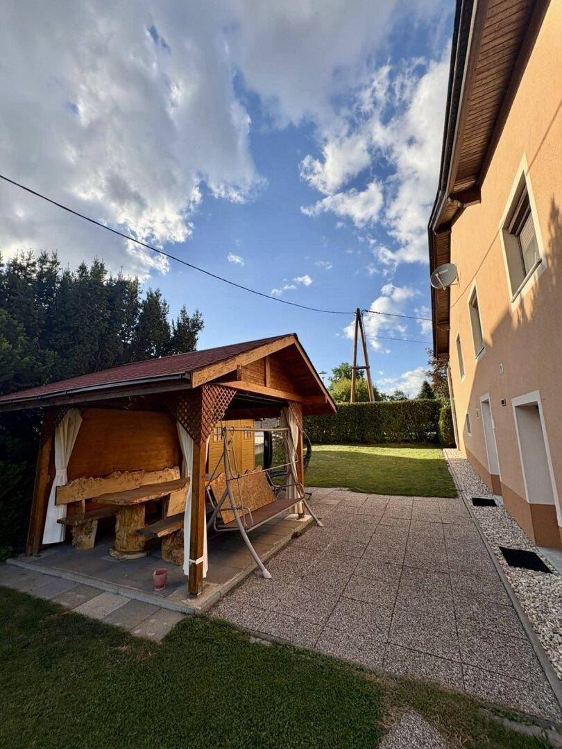 Seitenansicht des Hauses mit gepflastertem Bereich, Holzpavillon und weitläufigem Rasen.