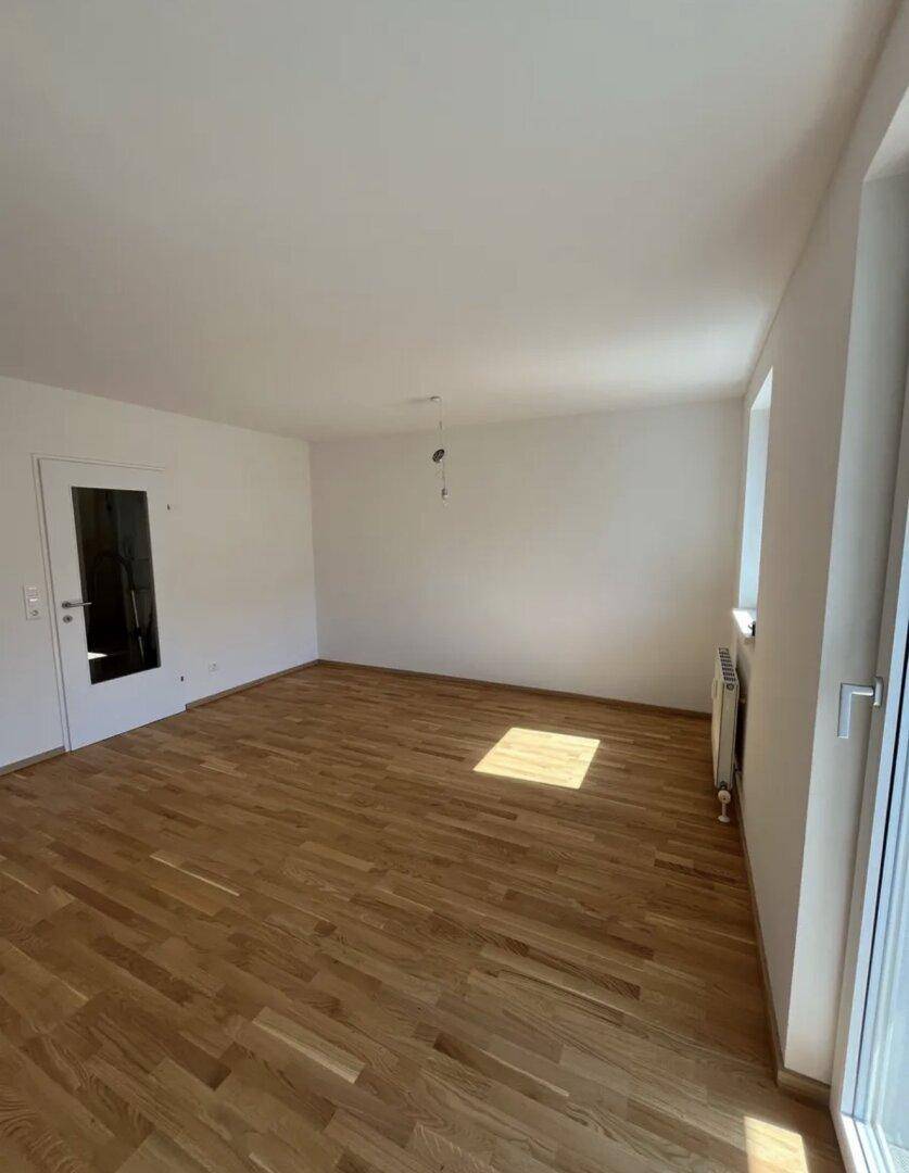Leeres Wohnzimmer mit hellem Parkettboden, weißen Wänden und viel Tageslicht durch große Fenster.