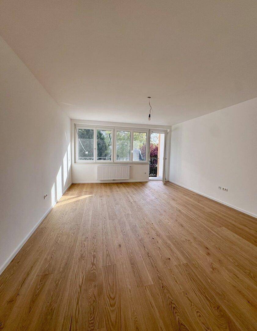 Helles Wohnzimmer mit Holzboden und direktem Zugang zu einem sonnigen Balkon mit Grünblick.