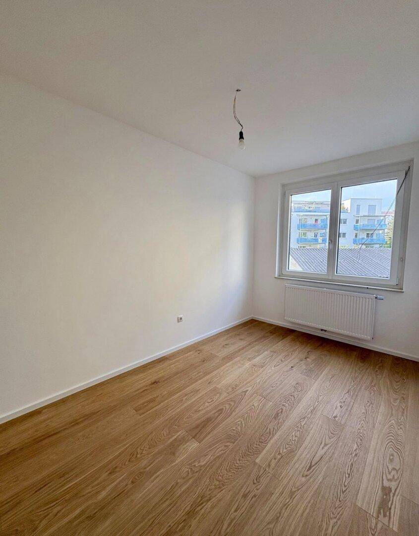 Lichtdurchflutetes Zimmer mit Holzboden und sauber gestrichenen Wänden, mit Blick ins Grüne.