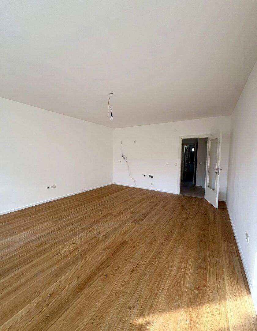 Leeres Zimmer mit Holzboden und weiß gestrichenen Wänden, mit offener Tür zum Flur.
