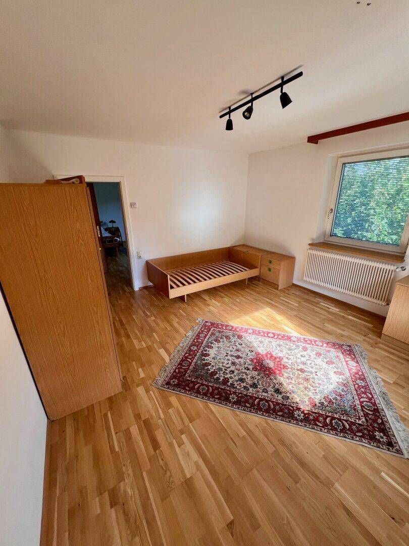 Schlafzimmer mit Holzboden, einem Bett, einem Teppich und einem Fenster mit Blick ins Grüne.